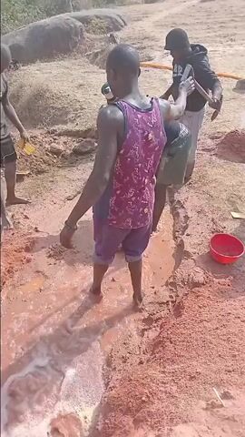 A group of men is engaged in outdoor work in a dry, open area with reddish-brown soil. In the foreground, a man in a purple patterned tank top and blue shorts stands barefoot in a muddy trench where water is actively flowing and bubbling. Two other men are bent over further along the muddy trench, also barefoot, seemingly working with the mud and water. To the right, another man in a black shirt and long pants uses a shovel to work with the dirt. Several other men are standing and observing the activity. One man in a red shirt stands with his hands on his head, while another in a brown jacket watches, and a man in a white tank top stands near a motorcycle. Red buckets and piles of reddish-brown sand or dirt are scattered on the ground. Yellow hoses are visible in the background. The scene is brightly lit by direct sunlight, casting clear shadows, indicating daytime. The environment appears arid or semi-arid. This activity is taking place in Agingi Zion, Nigeria.