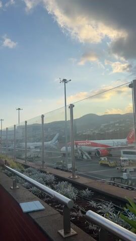 The video shows an airport tarmac at dusk. Several airplanes are parked at gates. One is an EasyJet airplane with its engines exposed. Airport vehicles such as luggage carts and a fire truck are also visible. There are a few people on the tarmac, likely airport personnel. The sky is cloudy with the sun setting behind a mountainous landscape. A glass barrier separates the tarmac from a walkway where the video is being filmed. Some plants are in the foreground.