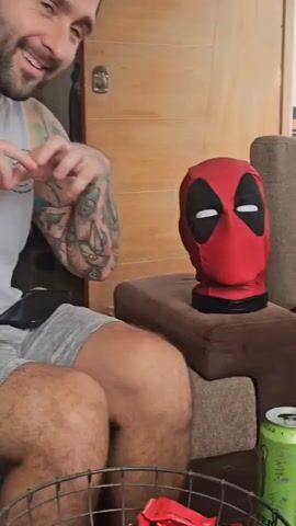 The video shows a man with dark hair, a beard, and tattoos sitting on a sofa indoors. He is wearing a gray tank top and gray shorts. He reaches out and touches a Deadpool head placed on a small ottoman next to him. He picks up his phone and begins to use it. A metal basket filled with small red candies is placed in front of him. A green can and a green bottle are also visible. A large wooden door is visible in the background.