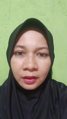 This is a close-up, front-facing video of a woman wearing a black hijab. She has light brown skin and is looking directly at the camera. The background is a plain, textured, light green wall. The lighting appears to be artificial and evenly distributed, with no indication of time of day or weather conditions. The woman’s facial expression is neutral. The video is short, with a timestamp of "00:00" to "00:04". There is no visible text in the video. The woman's lips are painted with a bright pink lipstick, and her eyebrows are neatly shaped. Her eyes are open, and she appears to be speaking or about to speak, as indicated by the slight parting of her lips in some frames. The overall impression is a personal video recording, possibly for social media or a personal message, given the close-up framing and direct address to the viewer.