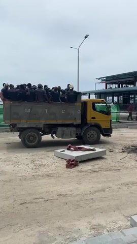 A yellow dump truck is parked on a dirt road, facing right. The truck bed is filled with approximately 20 people, all wearing dark blue shirts and some wearing yellow hard hats. They are all seated in the truck bed. A concrete slab with red straps lies on the ground in front of the truck. In the background, there is a building with a sign that reads "Coffee" and a street lamp. The sky is cloudy.
