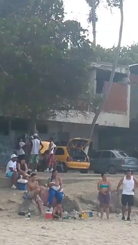 A group of people are gathered on a sandy area near a body of water. In the background, there is a yellow car with its trunk open, and several other parked cars. Behind the cars, there is a partially constructed building surrounded by trees. Some people are sitting on rocks, while others are standing. There are coolers, bags, and some bottles on the ground. The weather appears to be overcast.