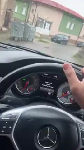 From the driver's seat of a Mercedes-Benz, a person grips the steering wheel as the vehicle idles on a wet street in a residential area. Through the rain-streaked windshield, several houses are visible, painted in light green, beige, and white. Several other vehicles, including an Audi A3, are parked along the street. Three metal garbage bins are located on the left. The car's dashboard displays system messages in a language other than English, and indicator lights are illuminated.