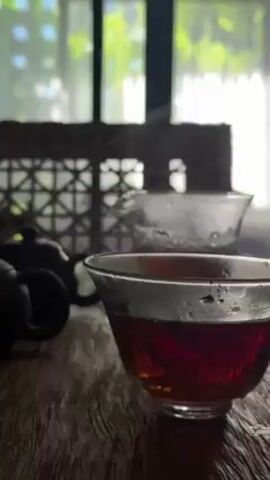 A tea ceremony is taking place on a dark wooden table. A glass cup is filled with a dark reddish-brown tea. Steam is rising from the cup. In the background, two dark teapots are sitting on the table. A small black teacup is visible in the lower left corner. A candle in a glass jar is also on the table, casting a warm light. A window with a patterned wicker screen is in the background, with sunlight filtering through the trees outside. The camera slowly pans across the scene from right to left.
