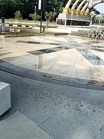 The video shows an exterior shot of an outdoor plaza with a large circular tiled pattern in the center. A bronze statue of a muscular human-like figure holding what looks like a large bone is prominently placed in the center of a water fountain. Behind the figure is another monument with a golden, claw-like design. Trees are visible in the background, and a small building with a red roof stands to the right of the figure. The camera pans upward to show the entirety of the statue and monuments. The sky is blue and the lighting suggests it's daytime.