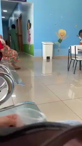 The video shows a waiting room, likely in a medical facility or clinic, decorated with colorful cartoon illustrations of children on the walls. A person is holding a clear plastic cup in their hand. The room has shiny tiled floors and contains rows of chairs, a small table, and a trash can. A few people can be seen in the background, one wearing a green uniform.