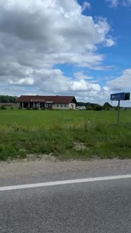 The video shows a POV shot from a vehicle driving on a paved road. The scenery includes a building with a red roof and green fields on the left side, followed by a church with a white facade and a graveyard. The video then transitions to show a modern white building with large windows and a sign above the entrance, followed by a shot of a parking lot with charging stations for electric vehicles. The sky is blue with scattered clouds, and there are several trees visible throughout the footage. The road has white markings.