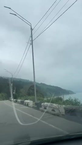 The video shows a view from inside a car driving on an asphalt road. The sky is overcast. There are utility poles and streetlights along the side of the road. A concrete barrier with arched cutouts runs along the side of the road overlooking a green hillside and a body of water, possibly the sea. A few cars are visible in the distance. The windshield of the car has some streaks on it.