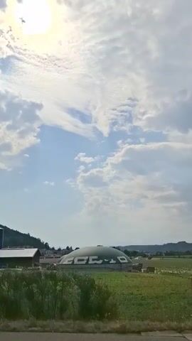 The video shows a rural landscape under a partly cloudy sky. The sun is visible in the upper left corner, casting a bright light. In the middle ground, there are several farm buildings, including a tall silo and a large, dome-shaped structure. The dome has the word "ECHO" written on it in large white letters. A field of green grass and vegetation stretches across the foreground. The overall impression is one of a working farm or industrial facility in a natural setting. The camera appears to be moving, possibly from a vehicle.