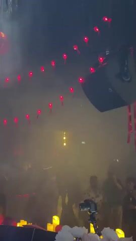 The media file captures a lively scene at a venue, likely a nightclub or event space, in Ban Bang Thao, Thailand. The setting is dark and filled with haze or smoke, illuminated by numerous red lanterns strung across the ceiling and a large red lantern hanging prominently. The atmosphere is festive, with people visible in the background, some appearing to be dancing or enjoying themselves.

In the foreground, several objects are visible. A DJ setup with turntables and controls is present, suggesting a musical performance is underway. A camera on a tripod is positioned to capture the event. The area around the DJ booth is decorated with a series of yellow, candle-like lights.

The people in the foreground are mostly silhouetted due to the lighting, but some figures are discernible. One person in the foreground appears to be wearing headphones and is positioned behind the DJ equipment, indicating they might be the DJ. Another person is visible with an arm raised in a celebratory gesture. The overall mood conveyed is energetic and celebratory, with the lighting and decorations contributing to a dynamic ambiance. The time of day is likely evening or night, consistent with the indoor, illuminated setting. No visible text is present in the media file.