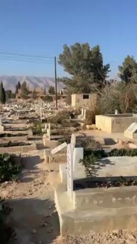 The video shows a graveyard with many headstones under a bright blue sky. The headstones are mostly white, and there are patches of green vegetation growing around them. Some of the graves are bordered by concrete. In the background, there are houses and trees, and a mountain range is visible in the distance. Power lines run across the sky. The graveyard appears to be well-maintained.