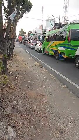 The video shows a congested road with a line of vehicles. The camera is positioned on the side of the road, looking down the street. In the foreground, there is a dirt path with some dry grass and debris. The road itself is paved and marked with a white line. Several cars and a green and yellow bus are visible in the traffic jam. The bus has "Abhiza Trans" written on its side. A white van is also present. Buildings and trees line the road, and there are utility poles and antennas in the background. The sky is overcast. The vehicles are stationary, indicating a traffic jam.