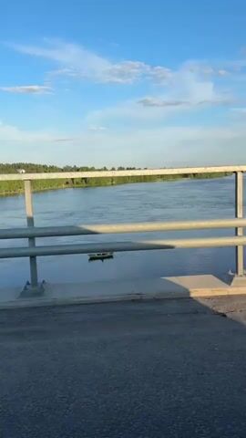 The video captures a serene view from a bridge crossing a wide river under a bright blue sky with scattered white clouds. The camera moves forward, suggesting a vehicle is crossing the bridge. In the foreground, a grey metal railing of the bridge is visible, with the road surface appearing as asphalt.

Beyond the railing, the calm, blue-grey water of the river stretches into the distance. On the far banks of the river, a continuous line of green trees forms a forested shoreline under the clear sky. A small, light-colored object, possibly a boat or buoy, can be faintly seen on the water's surface in the mid-ground. The lighting suggests it is either morning or late afternoon, with clear visibility and pleasant weather. The setting is a natural landscape with a prominent body of water and forested areas. No people or visible text are present in the video. The location is Imatra, Finland.