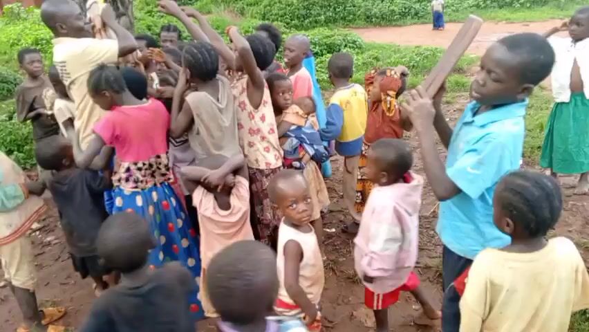 A group of approximately 20 children, mostly appearing to be of African descent, are gathered outside on a dirt patch in a wooded area. They are wearing casual clothing, with a variety of colors and patterns visible. An adult male, possibly a teacher or caregiver, is interacting with the children, gesturing with his hands while speaking. Some children are looking towards him, raising their hands. One child carries what looks like a wooden board on their head. There are trees and green foliage visible in the background, along with a small building in the distance. The scene seems lively and communal.