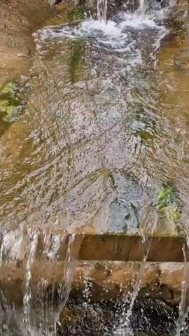 A close-up, downward-angle shot focuses on a small, man-made waterfall. The water cascades over a stone and concrete structure, collecting in a shallow channel below. The channel, lined with rocks and some sparse green vegetation, appears to be made of concrete or a similar material. The water is clear and creates ripples as it flows. The video progresses slightly upward, revealing more of the stone wall behind the waterfall and a hint of greenery and cloudy sky at the top of the frame. There are no people or animals visible in the video.