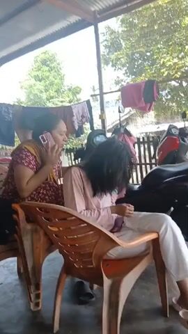 Two women are seated on chairs outdoors. The woman on the left is wearing a maroon and yellow patterned dress and is on a phone call. The woman on the right is wearing a light pink top and white pants. She is looking down at something in her lap, and her hair is covering her face. Clothes are hanging on a clothesline in the background, and there are motorcycles parked behind the women. The overall environment appears to be a residential area.