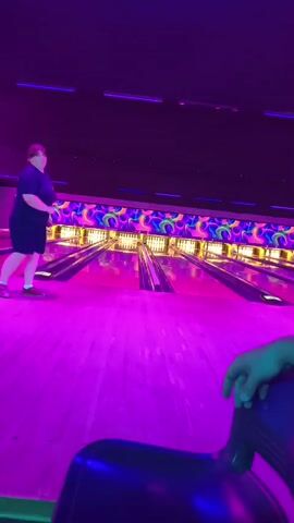 A person stands on a bright pink bowling lane, facing away from the camera. The bowling alley is dimly lit with purple and neon lights. The lanes are illuminated with neon lights. A person's hand rests on a dark blue chair in the foreground.