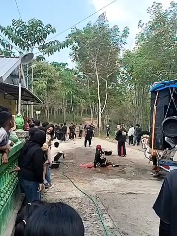 The video depicts an outdoor gathering of people. In the foreground, several individuals are dressed in black clothing. Some are standing and observing, while others are engaged in activities. One person is kneeling and appears to be washing something on the ground with a hose. Another person, also in black, is sitting on the ground next to a pile of pink material, which is wet. Behind them, a group of people, many in black attire, are standing and observing the scene. Some are holding what appear to be staffs or poles. In the background, there are trees and a dirt road. A large speaker system is visible on the right side of the frame, attached to a vehicle. The overall atmosphere appears to be a cultural or traditional event, possibly a performance or ceremony.