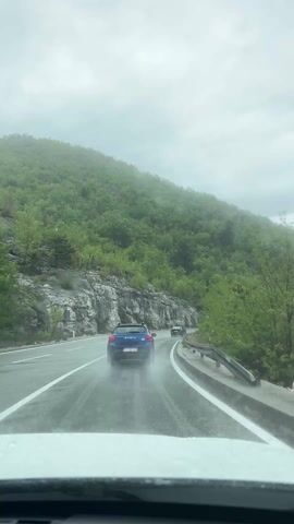The video captures a daytime view from inside a car driving on a wet, two-lane asphalt road through a mountainous region near Dobrsko selo, Montenegro. Raindrops are visible on the vehicle's windshield. A blue Dacia car, bearing the text "DACIA" on its rear and a Montenegrin license plate "ME BZ 270", is being followed, splashing water from its tires onto the wet road.

The road curves gently to the right, flanked by steep, verdant hillsides. On the left, there are rocky cliffs and cuttings, partially covered with green vegetation. On the right, a metal guardrail separates the road from a tree-filled slope. The sky is overcast, and the upper parts of the hills are partially obscured by mist or low-hanging clouds, indicating either ongoing rain or recent precipitation. The lush green foliage on the hills suggests a temperate climate. The scene conveys a sense of driving through a scenic, wet, and somewhat atmospheric natural landscape.
