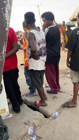 The video shows a group of young men standing on a street in what appears to be an urban area. There are about six or seven of them, and they are dressed casually in t-shirts, shorts, and sandals. Some are talking and gesturing. The surroundings include a partially open street drain, a food stall with an umbrella, and a building with signage for medical and general contract services. The overall impression is of a busy street scene with people socializing.