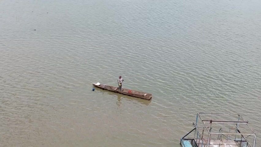 A man stands in a small boat on a body of water, possibly a lake or wide river. He is holding a long pole, perhaps using it to navigate or propel the boat. There is a bucket and a white sheet of material in the boat. Another structure is partially visible at the bottom right of the frame, possibly a platform or part of another boat. The water is calm with small ripples, and the sky is overcast.