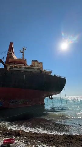The video shows a large, rusty shipwreck grounded on a rocky shore under a clear blue sky with a bright sun. The ship's hull is painted red and black, with some graffiti visible on the lower portion. The deck of the ship is weathered and appears abandoned. Small waves are breaking on the shore, which is littered with seaweed and white pebbles. In the foreground, two red plastic objects, possibly buckets or shovels, are visible on the pebbles. A few people are scattered on the rocky shore near the bow of the ship, some appearing to be sitting or standing. The camera pans slightly across the scene, capturing the vastness of the sky, the sun's glare, and the immensity of the beached vessel.