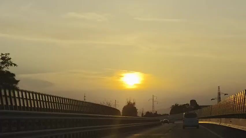 The video shows a view from a vehicle traveling on a highway at sunset. The sun is visible in the sky, appearing as a bright orb with some clouds around it. The sky is a warm yellow and orange hue, indicating the time of day. Guardrails are visible on both sides of the highway. Several other vehicles, including cars and vans, can be seen on the road ahead, some in the distance and some closer. Electrical pylons and trees are visible in the background. The lighting suggests it is either sunrise or sunset, with the sun low in the sky.