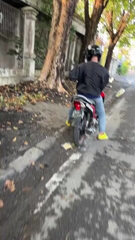 The video shows a person riding a motorcycle down a street. The person is wearing a black jacket, blue jeans, a helmet, and yellow shoes. The motorcycle is red and white. The street is paved with asphalt and has a sidewalk on the left side. There are trees on the left side of the street and a fence behind the trees. The weather appears to be overcast.