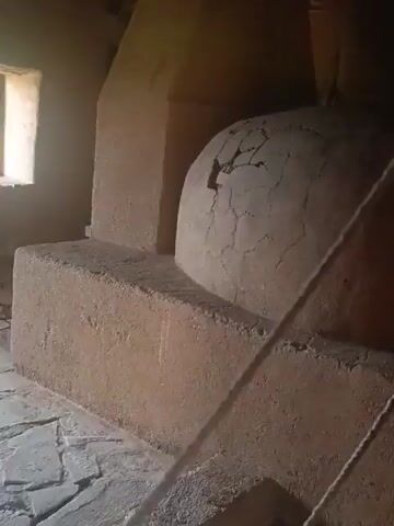 This media file shows the interior of an old, traditional building with a stone floor and earth-toned walls. The most prominent object is a large, dome-shaped oven, likely for baking bread or other food. It appears to be made of clay or a similar material and shows signs of age and use, with cracks visible on its surface.

To the left of the oven, there is a rectangular structure, possibly a hearth or a base for the oven. Further to the left, a window with a grid pattern allows natural light to enter the room. The light creates a bright, almost hazy effect, suggesting it might be daytime. Another similar window is visible on the opposite wall, further back in the room, also with a gridded pane. A light fixture hangs from the wooden ceiling beams.

The floor is covered with irregularly shaped flagstones, giving the space a rustic and historic feel. There are no people visible in the footage. The overall impression is of a historical structure, perhaps a rural dwelling or a communal building, preserved in time. The setting suggests a location in a historical or rural area, consistent with the provided location context of Rozhen, Bulgaria, known for its historical sites and traditional architecture. The lighting and the weathered appearance of the oven contribute to a sense of quiet antiquity and functional history.