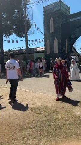 The video captures a sunny outdoor event, possibly a festival or fair, with a medieval theme. In the foreground, a person dressed in a colorful, striped robe and a crown walks towards the camera, holding a rolled-up fabric in their right hand. Their expression is cheerful. Behind them, other attendees are visible, some in casual wear, and one person in a white medieval-style gown. The background features a large, dark green structure resembling a castle tower with arched windows, and banners strung across the sky. The ground is a mix of dry grass and dirt. The overall atmosphere suggests a lively and festive gathering.