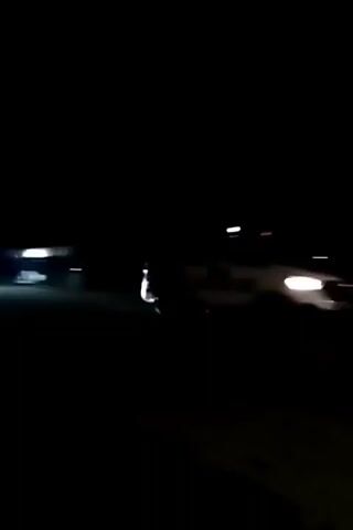 The video opens with a dark screen, then pans to a dimly lit road at night. A large vehicle, possibly a truck or an airplane, is visible in the distance on the left side of the road. Further down the road, there are lights from what appear to be buildings or structures. A traffic signal with a flashing red arrow indicating a sharp left turn is prominent on the right side of the road. Cars are driving on the road, their headlights illuminating the scene. The environment appears to be rural or suburban, with trees visible on the right side of the road.