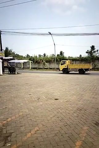 A yellow truck drives down a road next to a paved area. A person on a motorbike drives past the truck. Then, two people on a motorbike ride past the truck.