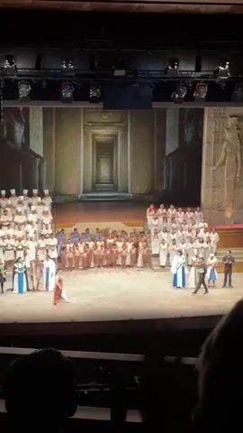 A video shows a wide shot of a stage during what appears to be a play or performance. The stage is set to resemble an Egyptian temple, with a large backdrop featuring stone structures and a long hallway receding into the distance. Onstage, there is a large cast of people in various costumes. Some are dressed in white robes and headwear, others in tan or brown clothing, and a few in more elaborate colorful costumes. 
The performers are arranged across the stage, some on raised platforms. At the front of the stage, several people appear to be taking bows, possibly in response to applause that can be heard in the background. The lighting is bright and theatrical, with spotlights highlighting the stage area. The video is recorded from the audience, with the back of someone's head visible at the bottom of the frame.