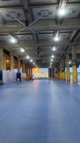 This video shows a wide shot of a large, open, covered area with a blue floor. The ceiling is concrete and features large industrial fans and numerous lights. There are many large concrete pillars supporting the ceiling. In the distance, through a large opening, a bright orange dome-shaped structure is visible, possibly a temple or monument. A few people are walking in the distance, and some are standing. Closer to the camera, on the left side of the frame, a group of people wearing orange robes, likely monks, are walking together. There are also some posters or displays on the left wall. On the right side of the frame, there are yellow pillars and some signage. The lighting suggests it is either dawn or dusk. The overall impression is that of a public space, possibly an airport terminal or a large gathering area.