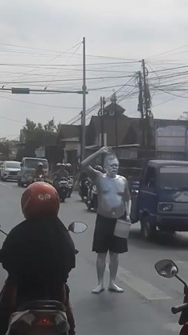 A man painted silver stands in a street with his arm raised. He is wearing black shorts and holding a bucket. Traffic, including motorcycles and a truck, moves around him. The scene appears to be in a town or city, with buildings and power lines visible.