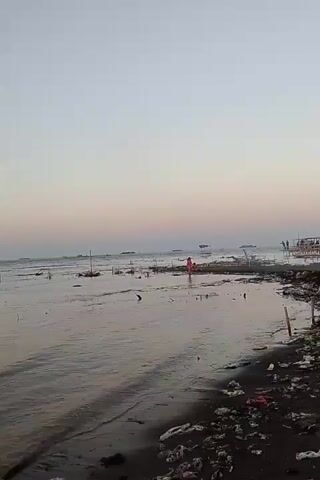 The video shows a wide, calm ocean with gentle waves lapping at a dark, debris-strewn beach. In the distance, a hazy horizon is visible where the sea meets a soft, gradient sky. Scattered across the water are several thin, vertical poles, some forming lines, suggesting a shallow area or a man-made structure such as a fish farm or a boundary. The beach itself is covered in litter, primarily plastic bags and other refuse, which are clearly visible along the shoreline. The overall impression is one of a natural landscape impacted by human waste, set against a serene, dusky sky.