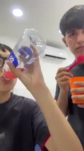 Two young people are indoors, likely in a kitchen or common area. A person on the left, wearing glasses and a black t-shirt, is drinking from a clear plastic bottle of "smartwater". The person on the right, also in a dark t-shirt, is holding an orange plastic cup and stirring its contents with a red plastic spoon. Behind them, a white tile wall is visible, as well as an air conditioning unit mounted high on the wall. A white appliance, possibly a refrigerator, is also partly visible. The lighting is bright and even. The subjects appear to be interacting with each other while looking at the camera.