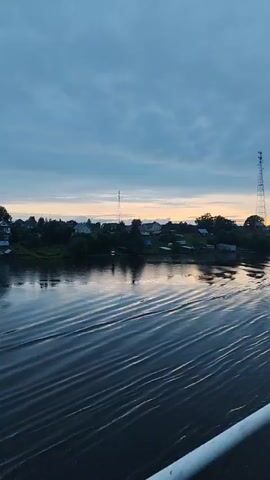 The video shows a wide river with ripples on its surface, reflecting the overcast sky and a distant shoreline. Along the shoreline, there is a cluster of houses and trees. The sky is a muted blue with streaks of lighter color near the horizon, suggesting either dawn or dusk. There are no people or animals visible in the frame. The camera appears to be moving, as evidenced by the waves creating a sense of forward motion. The overall mood is tranquil and serene.