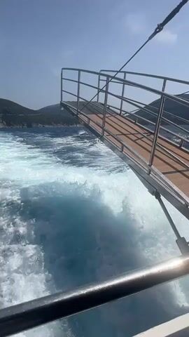 The video shows a close-up view of a ramp extending from a boat into the ocean. The water behind the boat is churning white with a visible wake. The sky is clear and blue, and hills covered in green vegetation can be seen in the background. The ramp appears to be made of wood with metal railings and is angled downwards towards the water. A rope is attached to the top of the ramp and extends upwards and out of frame. The camera is positioned low, looking out from the side of the boat, with parts of the boat's railing visible in the foreground.