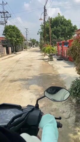 The video shows a first-person perspective of someone riding a motorcycle down a dirt road in a residential area. The road is lined with houses, trees, and utility poles. The sky is clear and sunny. In the foreground, the rider's hand, wearing a light blue sleeve, is visible on the handlebar. The motorcycle's dashboard is also partially visible. The rider is moving forward along the road, with houses and vegetation passing by on either side. The scene appears to be in a warm climate, given the foliage and sunny weather.