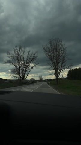 A stationary shot from inside a vehicle captures a straight asphalt road leading into the distance. On either side of the road are barren trees and green fields, while the sky above is covered in a heavy layer of dark, ominous clouds. The horizon is visible, revealing a distant landscape obscured by the atmospheric conditions. The black dashboard of the vehicle is visible at the bottom of the frame.
