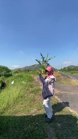 A person wearing a pink hijab and light-colored pants stands in a grassy area next to a paved road. They are holding a dark, pinstriped garment, possibly a shirt or jacket, and appear to be swinging it around their head. The background shows a clear blue sky, a hill in the distance, and some vegetation, including a banana tree. The sun is shining brightly, casting shadows.