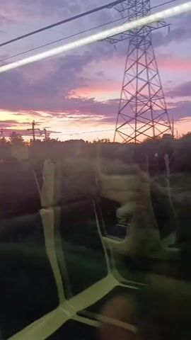This media file captures a view from a train, looking out a window at a rural landscape during a vibrant sunset. The sky is a dramatic display of purple and pink clouds, with streaks of orange near the horizon. Silhouetted against the colorful sky are trees, utility poles, and some distant buildings, suggesting a small town or village. The foreground of the image is dominated by the interior of the train carriage, with the reflections of seats visible on the glass. The seats appear to be empty, adding a sense of quiet observation to the scene. The overall mood is peaceful and contemplative, enhanced by the beautiful natural light and the gentle movement of the train. The time of day is clearly sunset. The location is identified as Shemodan, Russia. There is no visible text in the media file.