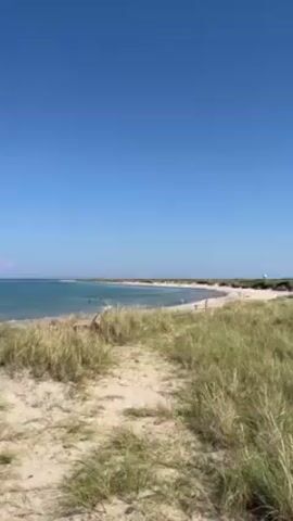 The video shows a sandy path leading towards a beach on a sunny day. The path is bordered by tall green grass. In the distance, a beach is visible with several people on it. The water is a light turquoise color, and the sky is a clear blue. There are a few white structures along the horizon line.