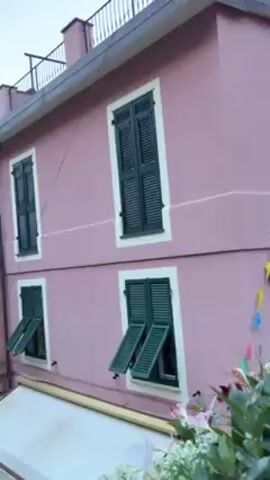 The video shows a narrow street in a European town, possibly in Italy. The buildings on either side are colorful, with a variety of pastel shades like pink, yellow, green, and blue. Many buildings have balconies and shutters, some of which are open. Laundry lines are strung between some of the buildings. The street slopes downwards toward the sea, which is visible in the distance under a clear blue sky. Several people are walking down the street, and a few small boats are parked alongside the road. A white awning stretches over a storefront in the foreground.