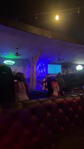 The video shows a dimly lit bar with neon lights and tropical-themed decor. Several people are present, some standing and some seated. The atmosphere appears to be lively, with music likely playing. A screen on the wall displays text that is partially visible. The camera pans across the scene, capturing different groups of people and aspects of the bar's interior.
