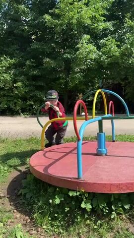 A young boy in a red plaid shirt and dark pants is playing on a merry-go-round in a park. He is holding onto the yellow bar of the merry-go-round and pushing off with his foot to make it spin. The merry-go-round is red and blue and is surrounded by green grass and trees. The boy is wearing a black baseball cap. He is spinning around and around. He is clearly enjoying himself. The sun is shining. It is a beautiful day.