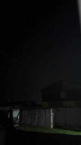 The video is a shaky, nighttime recording looking up at a dark sky. In the distance, two yellow lights appear to be moving. Below the lights, the roofline of a building is visible. In the foreground, a dark fence and some greenery can be seen.