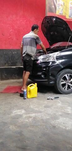A man in a grey and blue striped shirt and black shorts is working on the front of a black car with its hood open. He is standing on a red mat on a concrete floor. To his right, there is a yellow jug and a can of spray lubricant. Tools, including a wrench, are visible near the car. The environment appears to be a garage or workshop, with red walls on the left side of the frame.