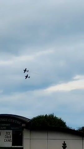 Two planes fly in formation leaving smoke trails against a cloudy sky. Below them is the top of a building with the words "THE HERITAGE CENTRE" and "SOUTHPORT CAFE" visible. A lamppost is also visible on the left side of the frame.